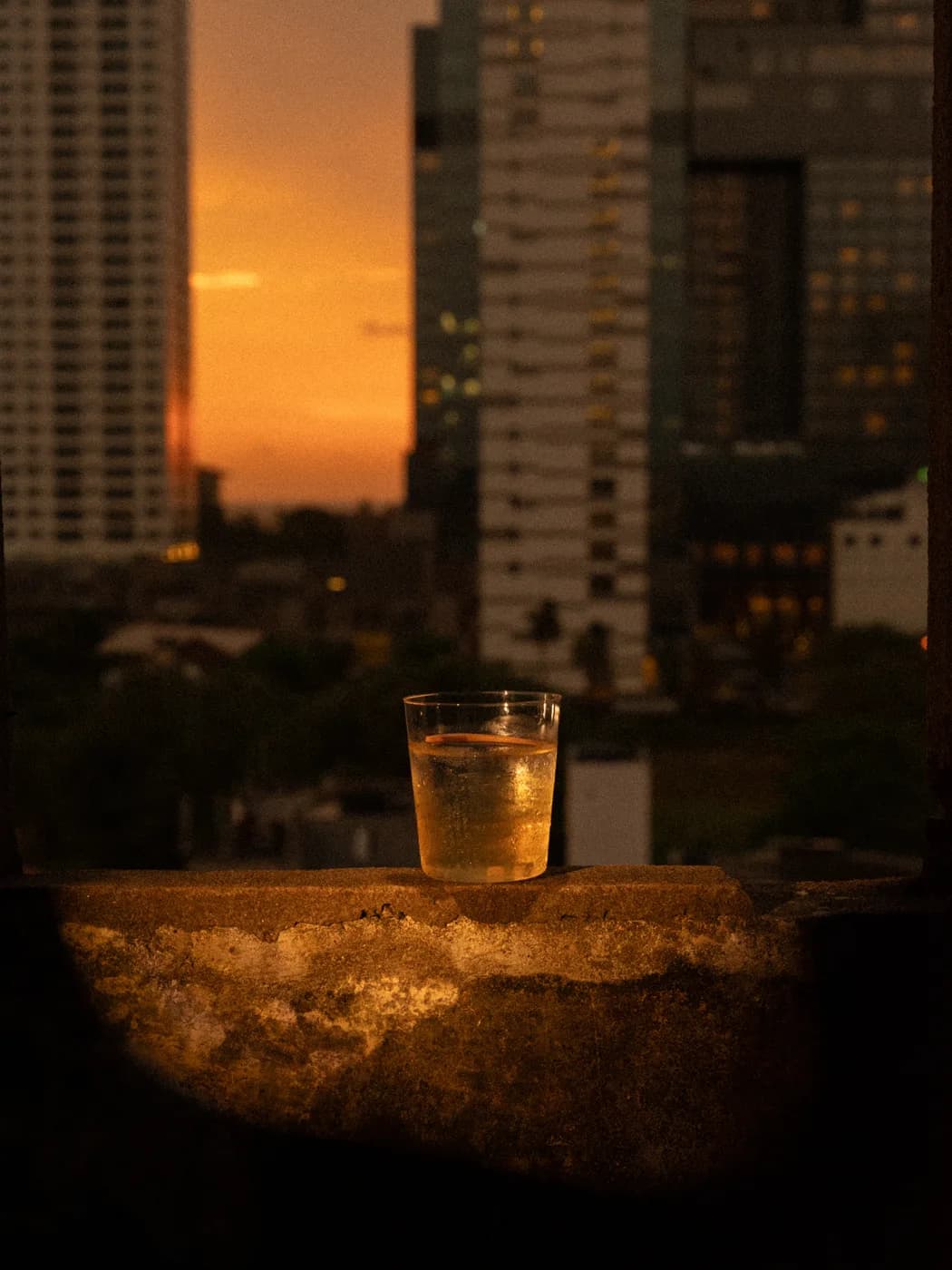 A glass on a concrete ledge with Colombo high-rises at sunset behind it