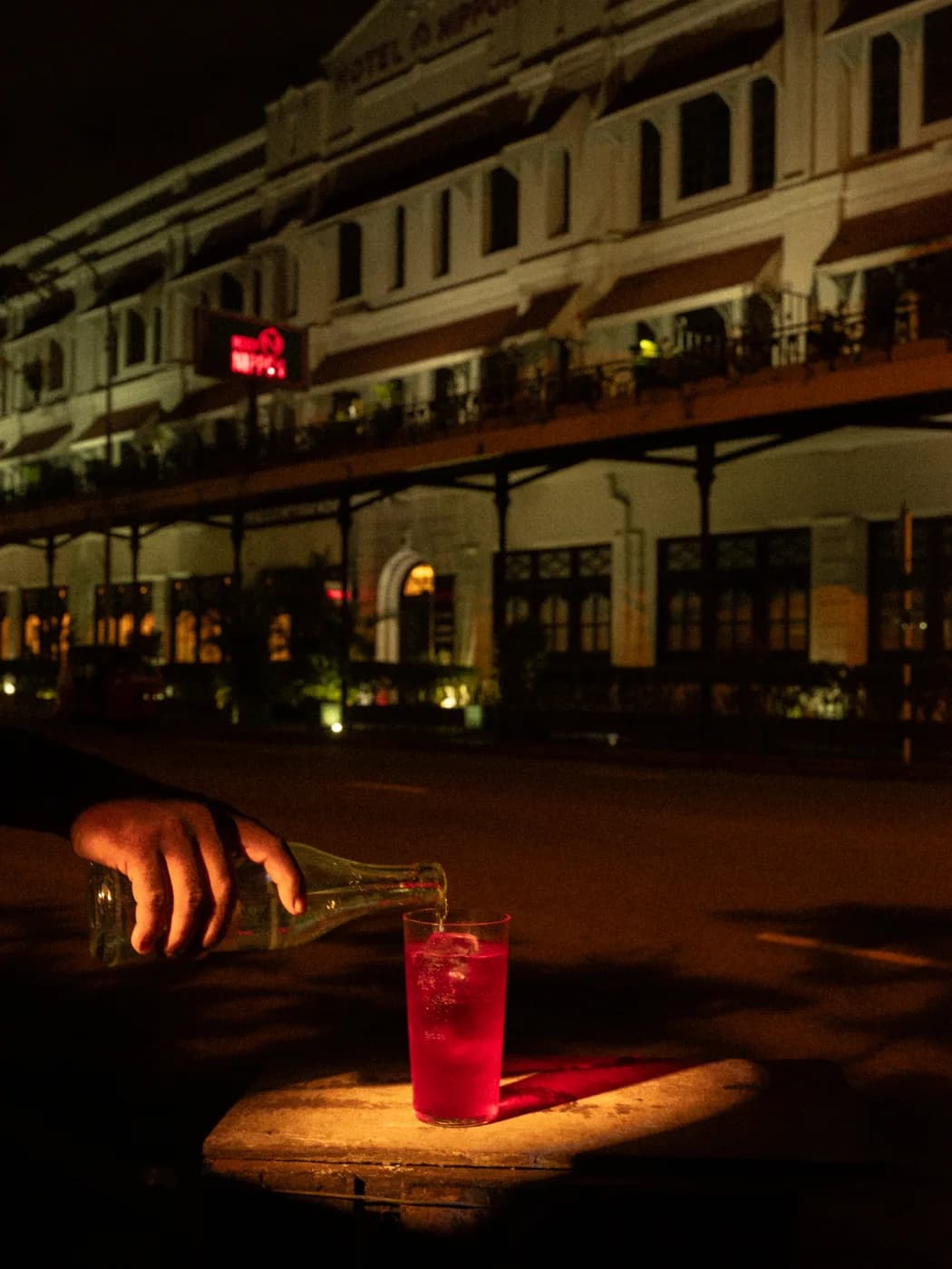 A hand pouring from a bottle into a red drink, a lit colonial facade behind